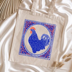 May include: A beige tote bag with a blue rooster illustration inside a red and blue patterned frame. The rooster is facing right and has a red comb.