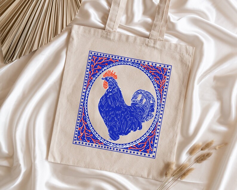 May include: A beige tote bag with a blue rooster illustration inside a red and blue patterned frame. The rooster is facing right and has a red comb.