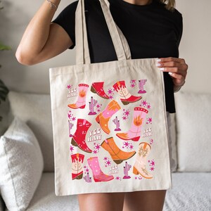 Könnte beinhalten: Eine beige Tragetasche mit einem bunten Muster aus Cowboystiefeln, Kakteen und Sternen. Der Text "Yee Haw" wird auf der Tasche wiederholt.