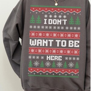 K&ouml;nnte beinhalten: Dunkelgraues Sweatshirt mit festlichem Design. Das Design enth&auml;lt den Text "I DON'T WANT TO BE HERE" in Wei&szlig;, zusammen mit gr&uuml;nen Weihnachtsb&auml;umen, roten Schneeflocken und wei&szlig;en Sternen. Ein gem&uuml;tlicher Weihnachts-Pullover.