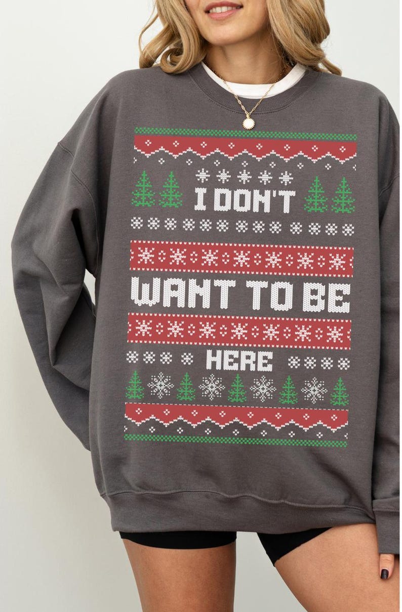 K&ouml;nnte beinhalten: Dunkelgraues Sweatshirt mit festlichem Design. Das Design enth&auml;lt den Text "I DON'T WANT TO BE HERE" in Wei&szlig;, zusammen mit gr&uuml;nen Weihnachtsb&auml;umen, roten Schneeflocken und wei&szlig;en Sternen. Ein gem&uuml;tlicher Weihnachts-Pullover.