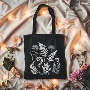 May include: A black tote bag with white botanical illustrations of ferns and other plants. The bag has a simple design and is perfect for carrying everyday essentials.