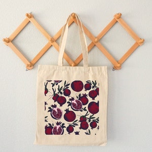 May include: A beige tote bag with a pattern of red pomegranates on a white background. The bag has a long handle and is hanging on a wooden rack.