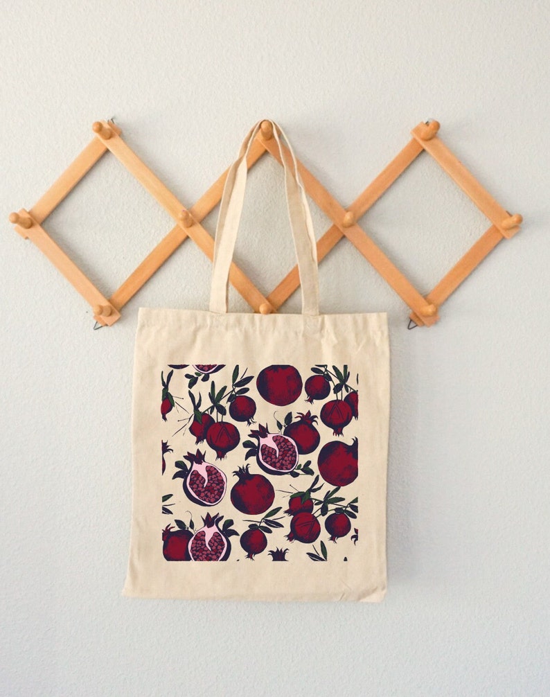 May include: A beige tote bag with a pattern of red pomegranates on a white background. The bag has a long handle and is hanging on a wooden rack.