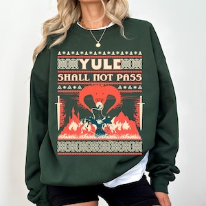 Peut inclure: Un pull de Noël vert avec un motif festif représentant un dragon, des épées et le texte "YULE SHALL NOT PASS".