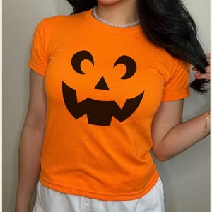 May include: Bright orange t-shirt with a black jack-o'-lantern design. The short-sleeved top has a round neckline. A comfortable and festive garment for Halloween.