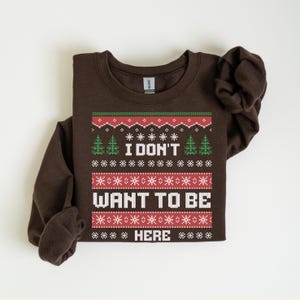 K&ouml;nnte beinhalten: Ein gefalteter brauner Pullover mit festlichem Design. Das Design enth&auml;lt den Text "I DON'T WANT TO BE HERE" in Wei&szlig;, mit roten und gr&uuml;nen Akzenten sowie Schneeflocken- und Tannenbaummustern. Der Pullover ist aus weichem Material.