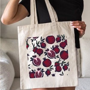 May include: A white canvas tote bag with a red pomegranate pattern. The bag has two long straps and is being held by a person.