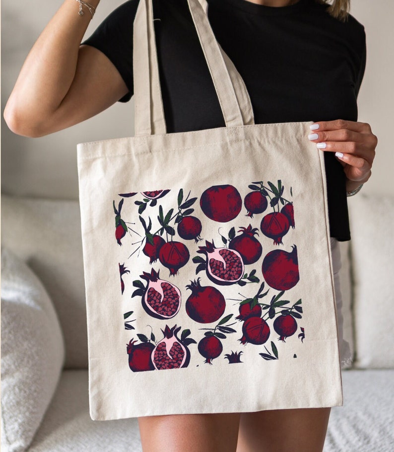 May include: A white canvas tote bag with a red pomegranate pattern. The bag has two long straps and is being held by a person.