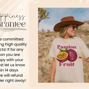 May include: A woman wearing a light beige t-shirt with a graphic of a passion fruit and the text "Passion Fruit". The woman is wearing a brown hat and is standing in a desert landscape.