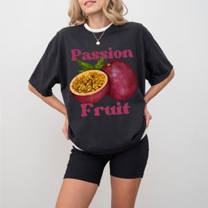 May include: A black t-shirt featuring a graphic of a passion fruit. The text "Passion Fruit" is printed in pink lettering on the shirt.