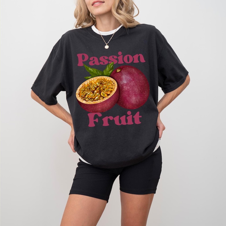 May include: A black t-shirt featuring a graphic of a passion fruit. The text "Passion Fruit" is printed in pink lettering on the shirt.