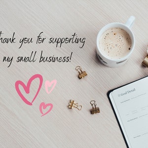 May include: A white coffee mug with a brown liquid inside, a gold watch, two gold paper clips, a pink heart, and a notebook with the text "Goal Detail" on the cover. The text "Thank you for supporting my small business!" is written on a light wood surface.