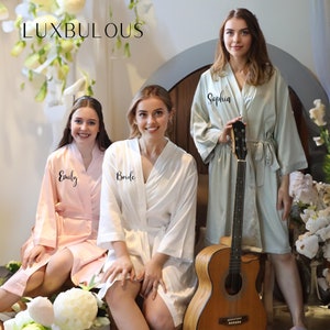 May include: Three women wearing satin robes in pink, white, and sage green. The robes have wide sleeves and are tied at the waist. The word "Bride" is on the white robe, and names are on the other robes. A guitar leans against one woman.