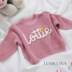 Puede incluir: Un suéter de punto rosa con el nombre "Lottie" bordado en hilo blanco con un pequeño acento de flor amarilla.