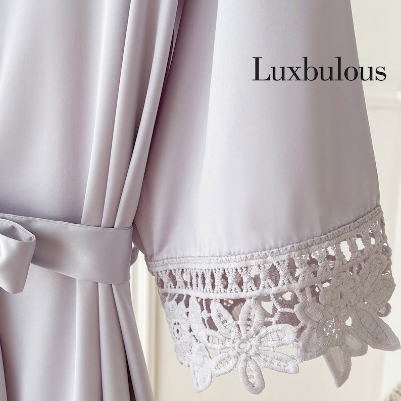 Op de afbeelding: Een lichtpaarse satijnen badjas met bijpassende ceintuur. De mouwboord heeft een delicate, witte bloemen kanten afwerking. Het woord "Luxbulous" is in het zwart rechtsboven in de afbeelding gedrukt.