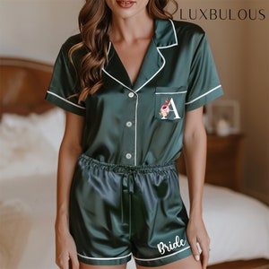 Op de afbeelding: Donkergroene satijnen pyjamaset met witte bies. De top met korte mouwen heeft een bloemenletter "A" op de zak. De shorts hebben "Bride" op de pijp gedrukt. De merknaam "LUXBULOUS" staat rechtsboven gedrukt.