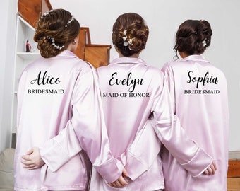 Puede incluir: Tres mujeres con batas de satén rosa claro a juego con texto negro en la espalda. El texto dice "Alice Bridesmaid", "Evelyn Maid of Honor" y "Sophia Bridesmaid".