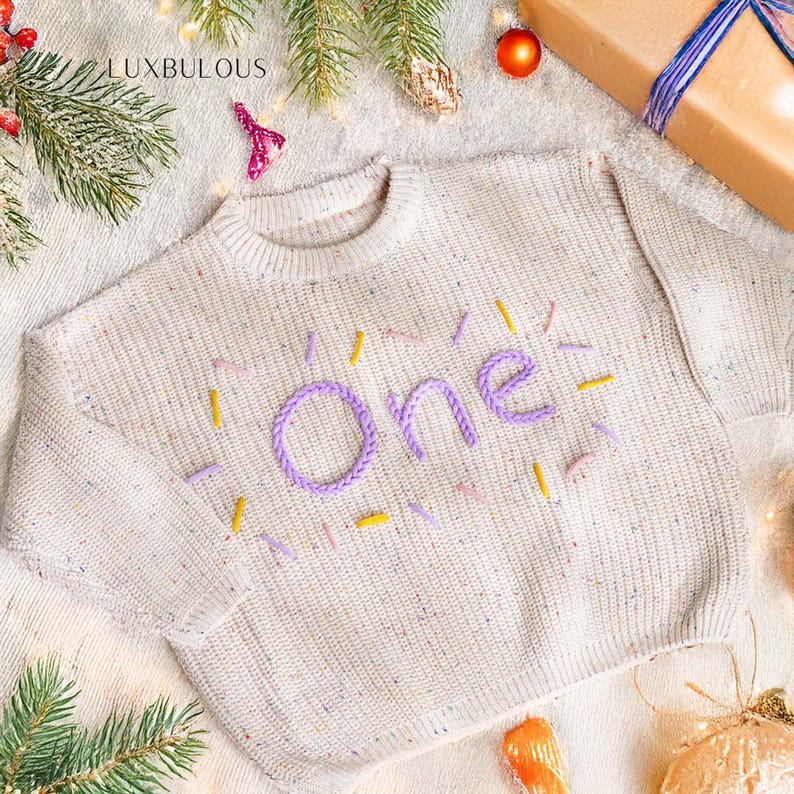 May include: A cream-colored sweater with the word "One" embroidered in purple, surrounded by yellow and purple accents. The sweater is speckled with various colors and has a round neckline. The word "LUXBULOUS" is in the top left corner.