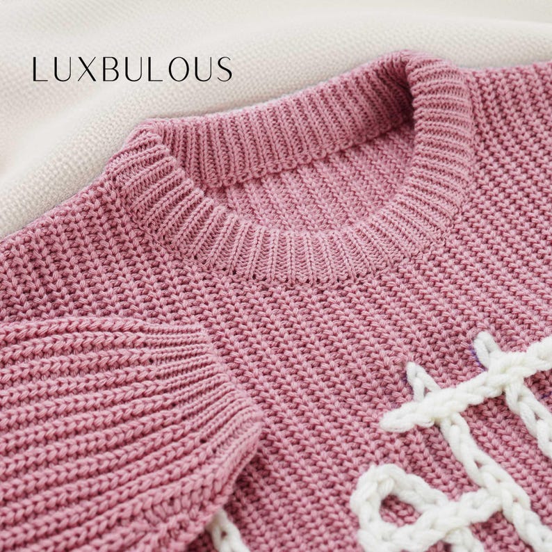 May include: A pink knitted jumper with a white knitted design. The jumper has a crew neck and long sleeves.