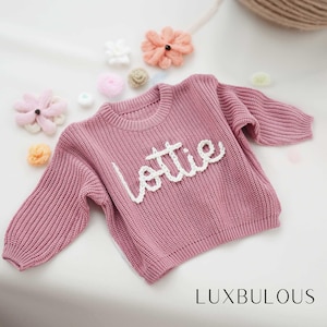 Könnte beinhalten: Ein rosa gestrickter Pullover mit dem Namen "Lottie" in weiß auf der Vorderseite gestickt.
