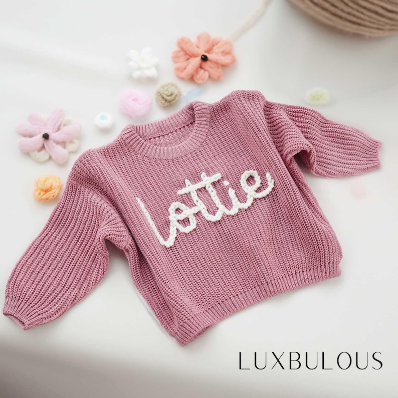 May include: A pink knitted jumper with the name "Lottie" embroidered in white on the front.