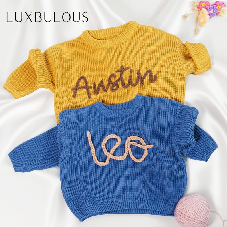 May include: Two knitted baby sweaters, one yellow with the name "Austin" embroidered in brown yarn and one blue with the name "Leo" embroidered in brown yarn.