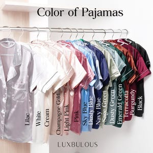 Op de afbeelding: Een verscheidenheid aan satijnen pyjamashirts in verschillende kleuren hangt aan een kledingrek. Kleuren zijn lila, wit, cr&egrave;me, champagne goud, lichtroze, roze, hemelsblauw, stoffig blauw, marineblauw, saliegroen, stoffig groen, smaragdgroen, terracotta, bordeauxrood en zwart. De tekst luidt "Color of Pajamas".