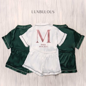 Op de afbeelding: Drie sets satijnen pyjama's. E&eacute;n witte set met een grote initiaal en de naam "Madelyn" en de datum "18.06.2024" op de achterkant gedrukt. Twee donkergroene sets staan aan weerszijden van de witte set. Het woord "LUXBULOUS" staat bovenaan.