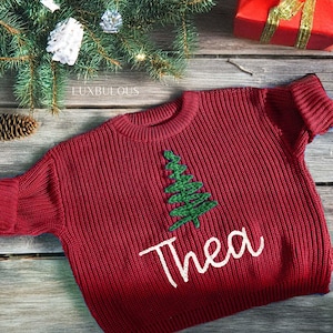 May include: A red knit sweater with a green embroidered Christmas tree and the name "Thea" in white script. The sweater is displayed on a wooden surface with Christmas decorations, including a gift box and pine needles.