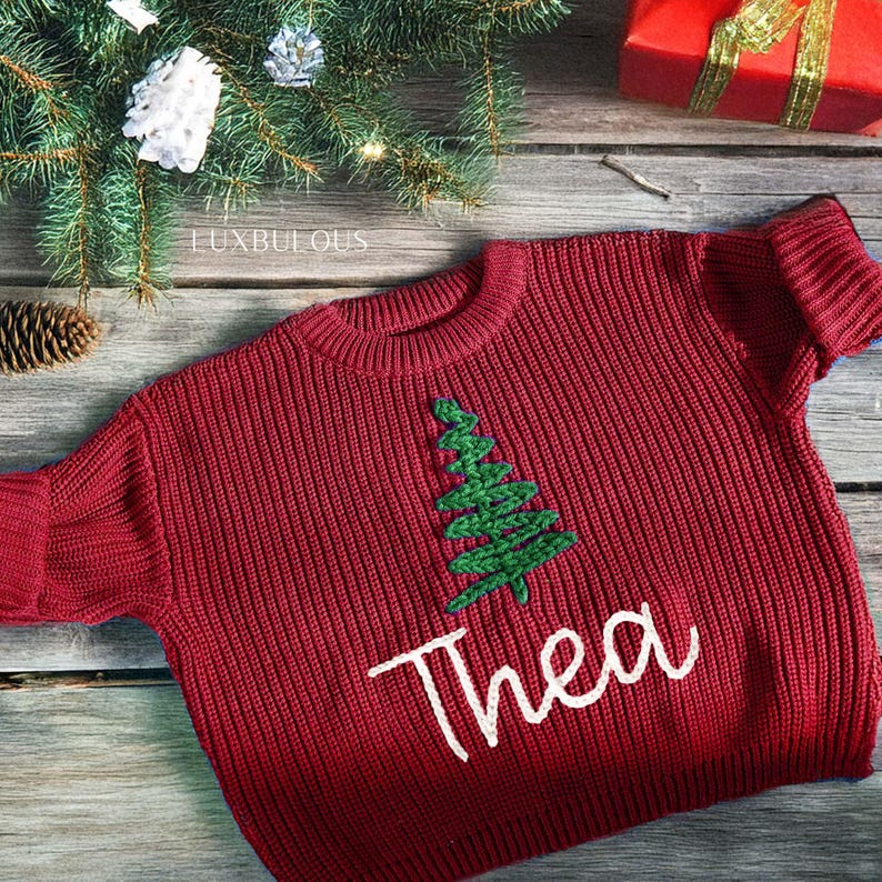 May include: A red knit sweater with a green embroidered Christmas tree and the name "Thea" in white script. The sweater is displayed on a wooden surface with Christmas decorations, including a gift box and pine needles.