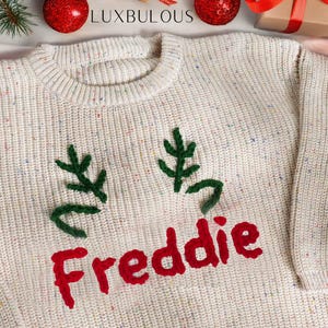 May include: Cream-colored sweater with green embroidered antlers and the name "Freddie" in red. The sweater has a speckled pattern and a ribbed crew neck. The word "LUXBULOUS" is at the top.
