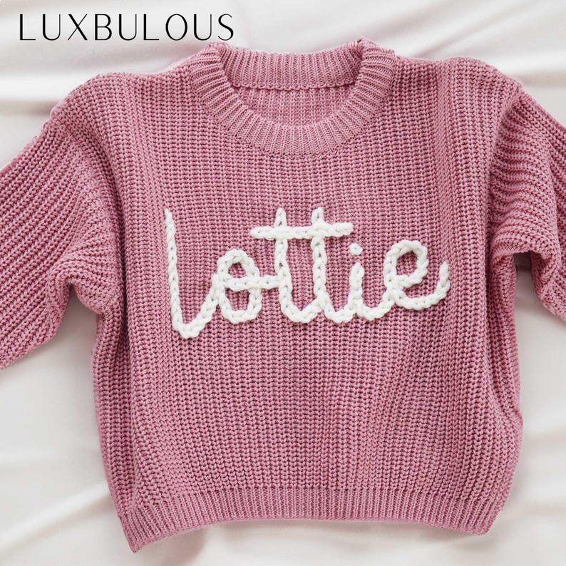 May include: A pink knitted jumper with the name "Lottie" embroidered in white yarn.
