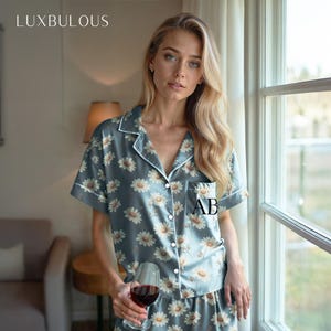 Può includere: Pigiama blu con motivo floreale a margherite. La maglia a maniche corte ha un colletto a revers e una tasca con le iniziali "AB". La parola "LUXBULOUS" è in alto a sinistra.
