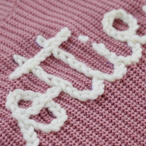 May include: Close-up of a pink knitted fabric with the word "LOVE" embroidered in white yarn.