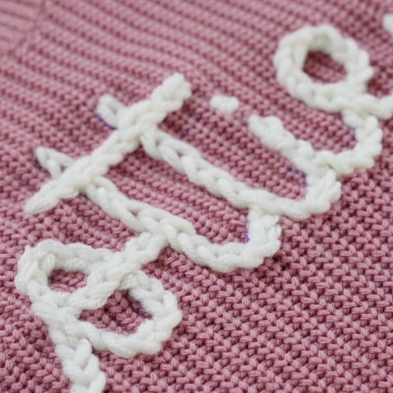 May include: Close-up of a pink knitted fabric with the word "LOVE" embroidered in white yarn.