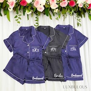Op de afbeelding: Drie sets satijnen pyjama's in marineblauw en zwart, met witte biezen. Elke set bevat een button-up shirt met korte mouwen en bijpassende shorts. De marine sets zijn geborduurd met initialen en het woord "Bridesmaid", terwijl de zwarte set "Brides" zegt.