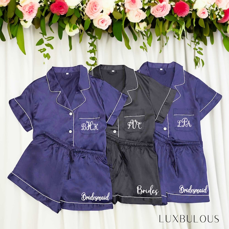 Op de afbeelding: Drie sets satijnen pyjama's in marineblauw en zwart, met witte biezen. Elke set bevat een button-up shirt met korte mouwen en bijpassende shorts. De marine sets zijn geborduurd met initialen en het woord "Bridesmaid", terwijl de zwarte set "Brides" zegt.