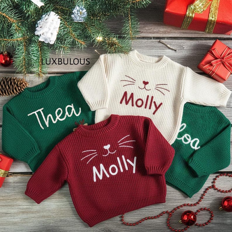 May include: Four children's sweaters in green, cream, and burgundy, with the names "Thea", "Molly", and "Loa" embroidered in white. The cream and burgundy sweaters feature a cat face design. Christmas-themed with a festive background.