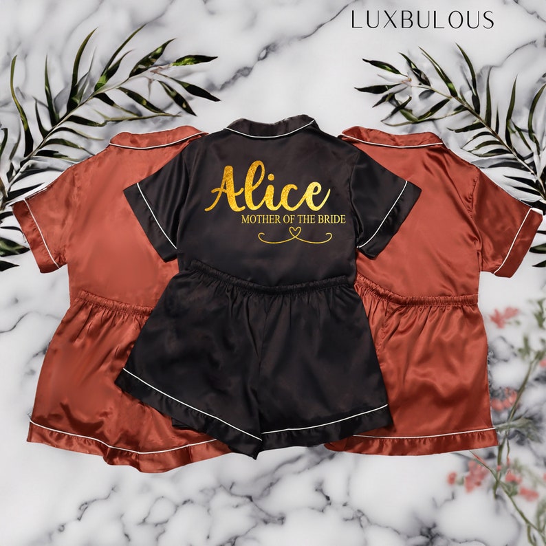 Op de afbeelding: Satijnen pyjamasets in roest en zwart. De zwarte set heeft de naam "Alice" en "Mother of the Bride" in gouden letters. Elke set bevat een top met korte mouwen en een short met witte bies. De sets worden gepresenteerd op een marmeren oppervlak.