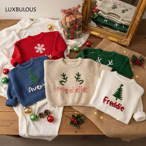 May include: A collection of children's Christmas sweaters in various colors, including red, blue, green, and white. Each sweater features festive embroidered designs such as snowflakes, Christmas trees, and reindeer antlers, with personalized names. The brand name "LUXBULOUS" is visible.