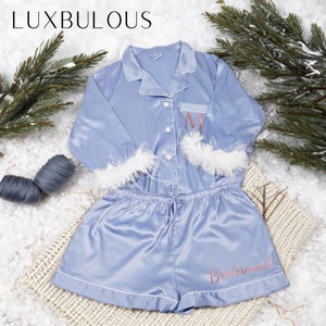 Könnte beinhalten: Ein hellblauer Satin-Pyjama-Set mit weißen Verzierungen. Das Oberteil hat lange Ärmel mit weißen Federverzierungen an den Manschetten. Die Shorts haben einen Kordelzugbund und das Wort "Brautjungfer" ist auf dem Bein gestickt. Das Oberteil hat einen Knopfleisten vorne und eine Tasche mit dem Buchstaben "M" darauf gestickt.