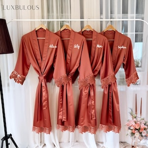 May include: Four terracotta-coloured satin robes with lace trim hang on a rail. Each robe has a matching belt and the names "Alice", "Lily", "Mia", and "Sophia" embroidered on the front. The robes are displayed in front of a white curtain.