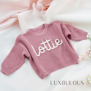 May include: A pink knitted sweater with the name "Lottie" embroidered in white yarn. The sweater is made by Luxbulous.