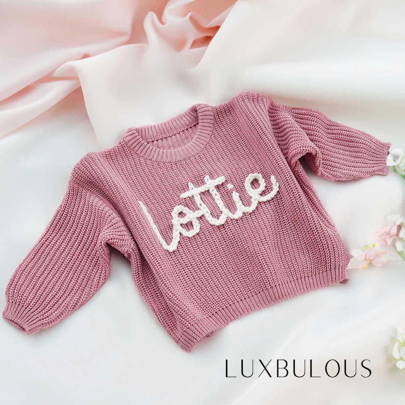 May include: A pink knitted sweater with the name "Lottie" embroidered in white yarn. The sweater is made by Luxbulous.