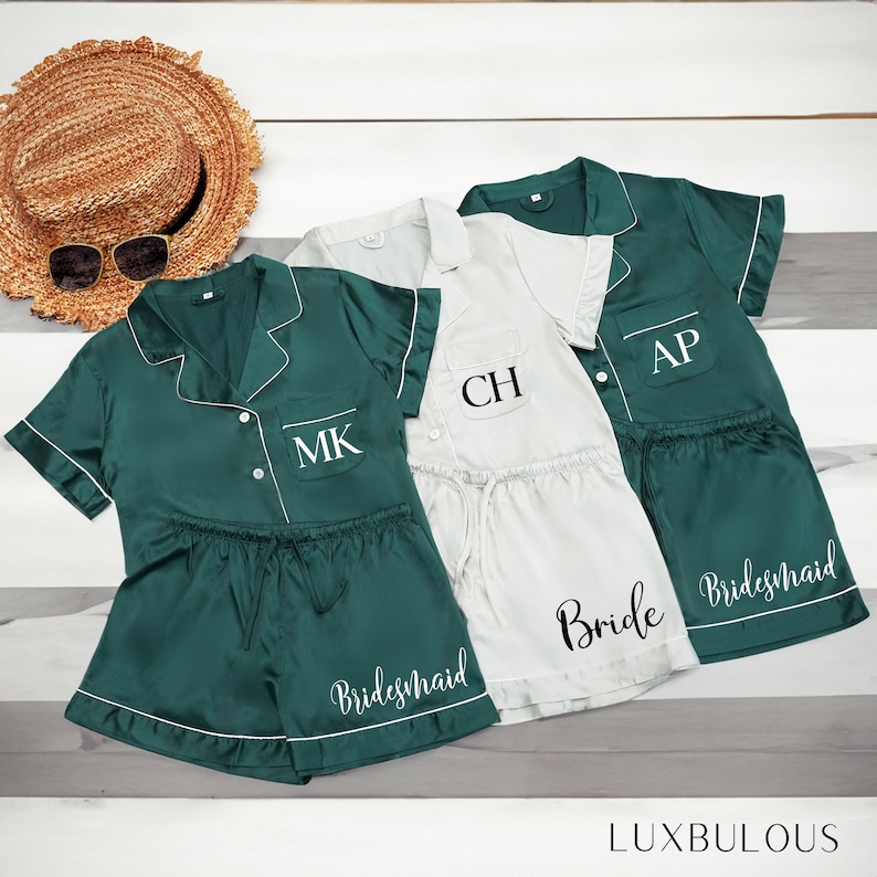 Op de afbeelding: Drie sets satijnen pyjama's in smaragdgroen en lichtgrijs, met witte biezen. De groene sets hebben de initialen "MK" en "AP" en het woord "Bridesmaid". De grijze set heeft de initialen "CH" en het woord "Bride". Een strohoed en zonnebril staan op de achtergrond.