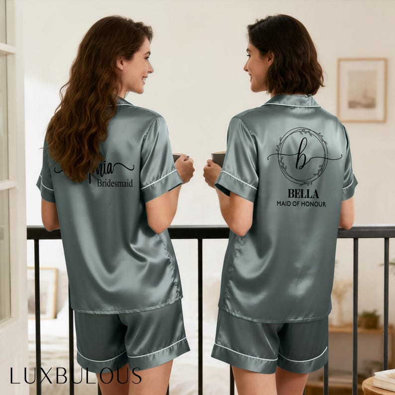 Op de afbeelding: Twee vrouwen in bijpassende grijze satijnen pyjamasets met witte bies. E&eacute;n set is gelabeld "Mia, Bridesmaid" en de andere "Bella, Maid of Honour". De pyjama's hebben korte mouwen en shorts. De merknaam "LUXBULOUS" staat onderaan.