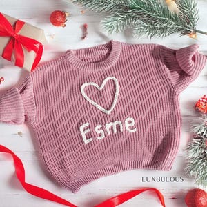 May include: A dusty rose-colored knit sweater with a white heart and the name "Esme" embroidered on the front. The sweater is surrounded by Christmas decorations, including a wrapped gift with a red bow and red ornaments.