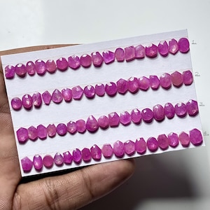 Może przedstawiać: Rzędy fasetowanych, różowych kamieni szlachetnych w różnych kształtach, w tym owalnych, łezkowych i sześciokątnych. Kamienie szlachetne są ułożone na białej karcie. Kamienie szlachetne mają żywy różowy kolor.