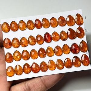 May include: A collection of faceted, teardrop-shaped gemstones in shades of orange and amber. The gems are arranged in rows on a white card, showcasing their varying sizes and internal structures. The gems are likely for jewelry making.